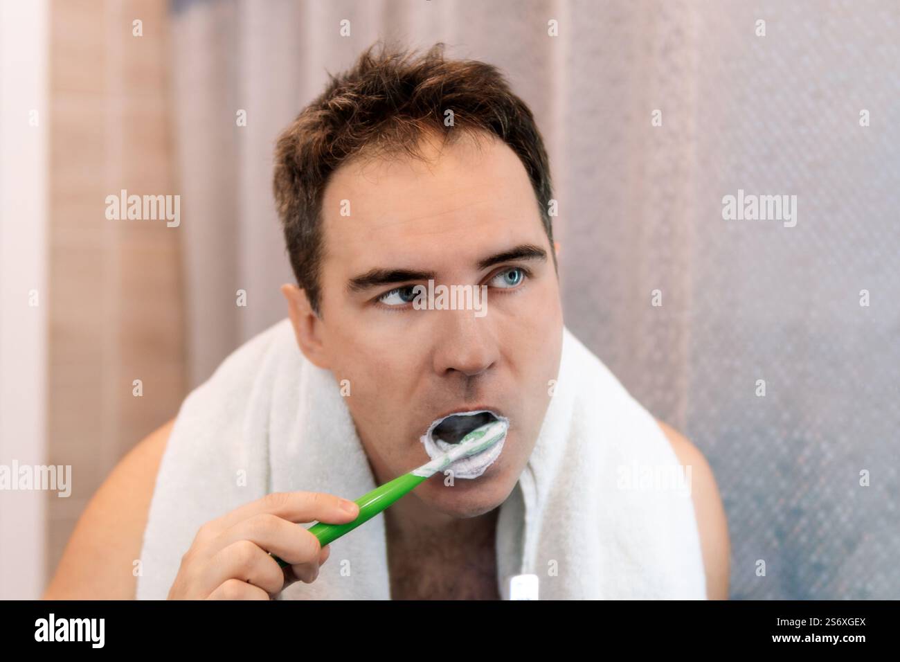 Hübscher junger bärtiger attraktiver kaukasischer Mann mit weißem Frottee-Bademantel lächelt und blickt auf seine Spiegelung im Badezimmerspiegel, während er Zier putzt Stockfoto