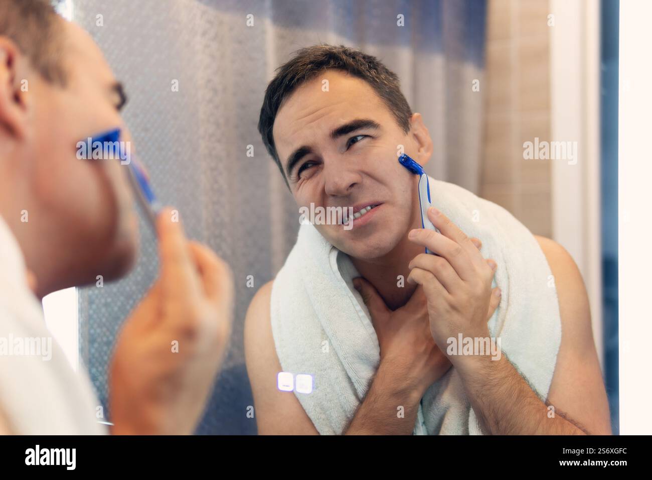 Ein Mann rasiert sein Gesicht ohne Creme oder Schaum und erlebt Schmerzen und Leiden. Trauriger Mann rasiert sich morgens vor einem Spiegel. Ungesunde Rasur von Stockfoto
