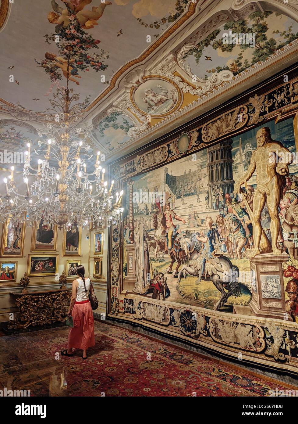 Ein üppiger Museumsraum mit kunstvollen Wandteppichen, vergoldetem Dekor und einem großen Kronleuchter, während ein Besucher in einem fließenden Rock die Kunst und Umgebung bewundert. Stockfoto