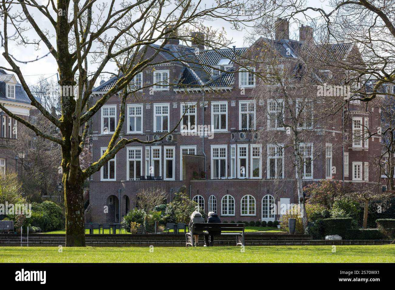 Gebäude in der Nähe des Vondelparks mit 2 älteren Menschen auf einer Bank im grünen Gras am Wasser im Winter, Amsterdam, Niederlande. 27. März 2023. Stockfoto