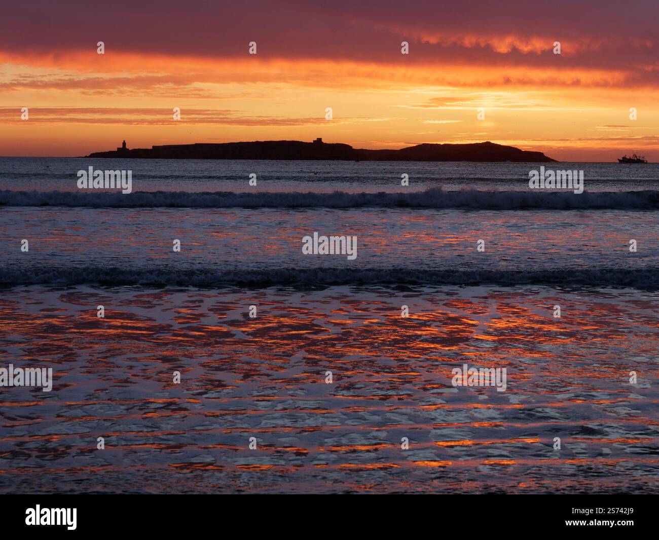 Dramatische dunkle Wolken bei Sonnenuntergang über einer Insel vor der Küste von Essaouira, während sich der feurige Himmel im Wasser spiegelt, Marokko. Januar 2025 Stockfoto