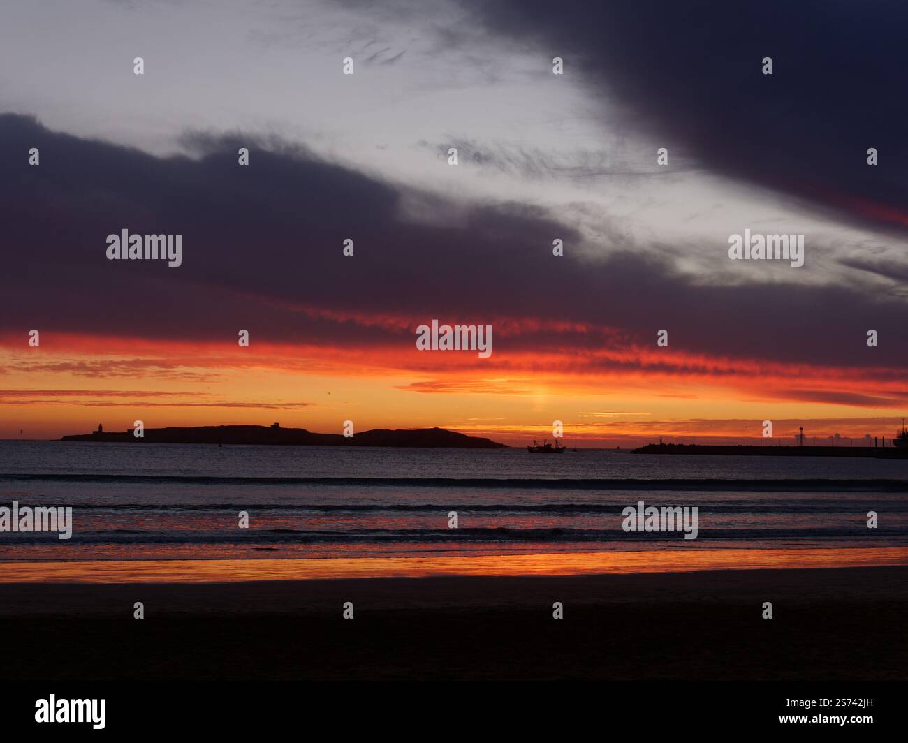 Dramatische dunkle Wolken und roter Himmel spiegeln sich im Wasser bei Sonnenuntergang über einer Insel vor der Küste von Essaouira, Marokko. Januar 2025 Stockfoto