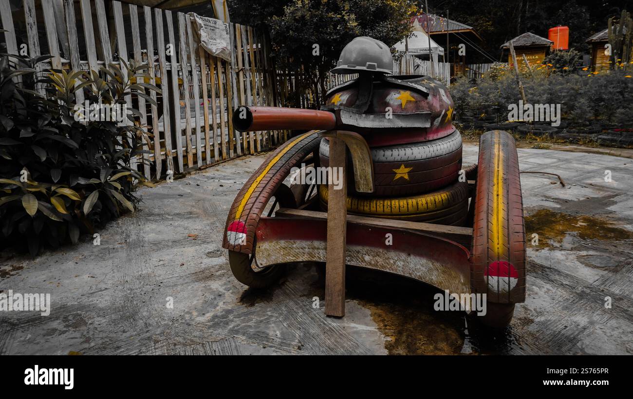 Eine skurrile Skulptur eines Panzers aus bunt bemalten Reifen und Holz, mit einem Helm oben, in einem Außenbereich mit Pflanzen und einem Zaun. Stockfoto