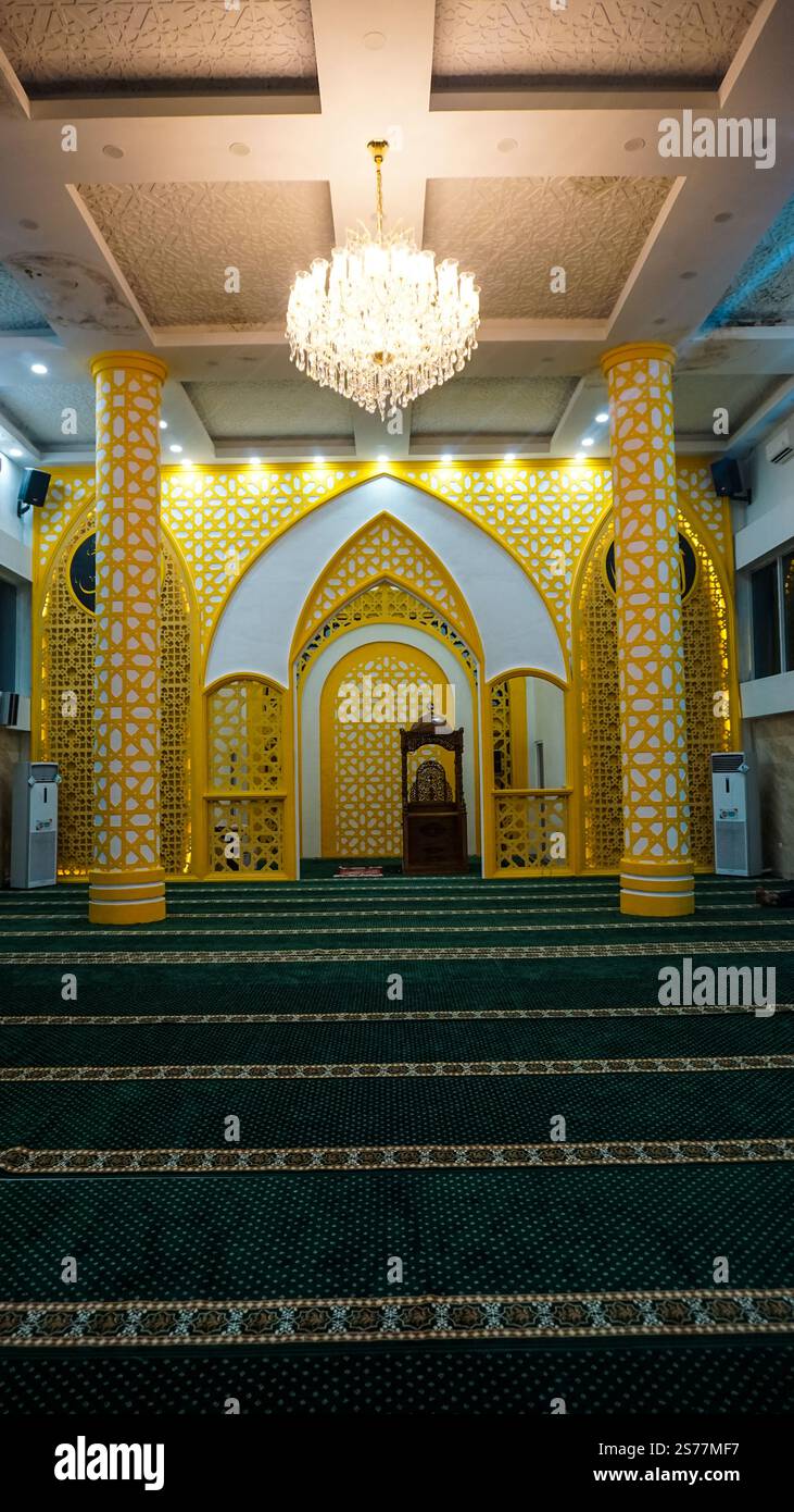 Ein wunderschön gestaltetes Moscheezimmer mit komplexen gelben und weißen Mustern, hohen Säulen und einem Kronleuchter. Der Gebetsbereich ist mit geschmückt Stockfoto