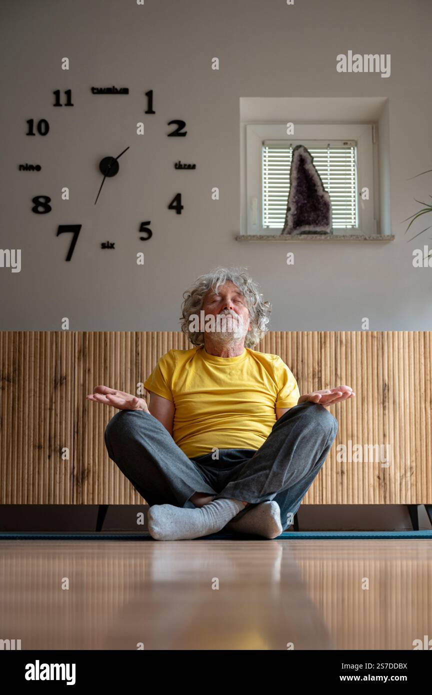 Ein älterer Mann in einem gelben Hemd sitzt in Lotus-Yoga-Position auf dem Boden und meditiert, mit einer modernen Uhr und Amethyst-Kristall im Hintergrund. Stockfoto