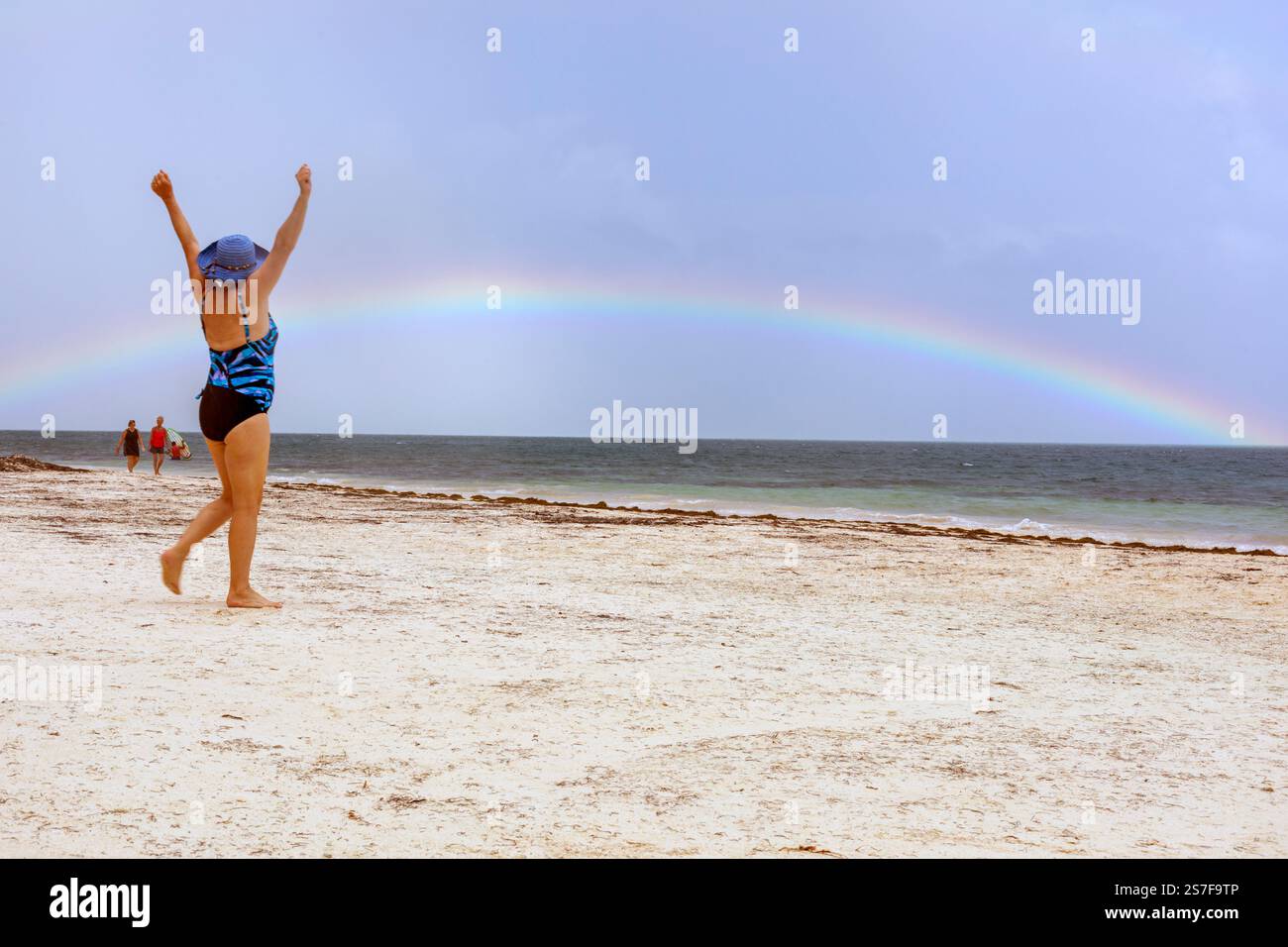 Mexiko, Puerto Morelos, Strandszene mit einer Frau, die ihre Arme vor Freude über dem Anblick eines Regenbogens über dem Wasser streckt Stockfoto