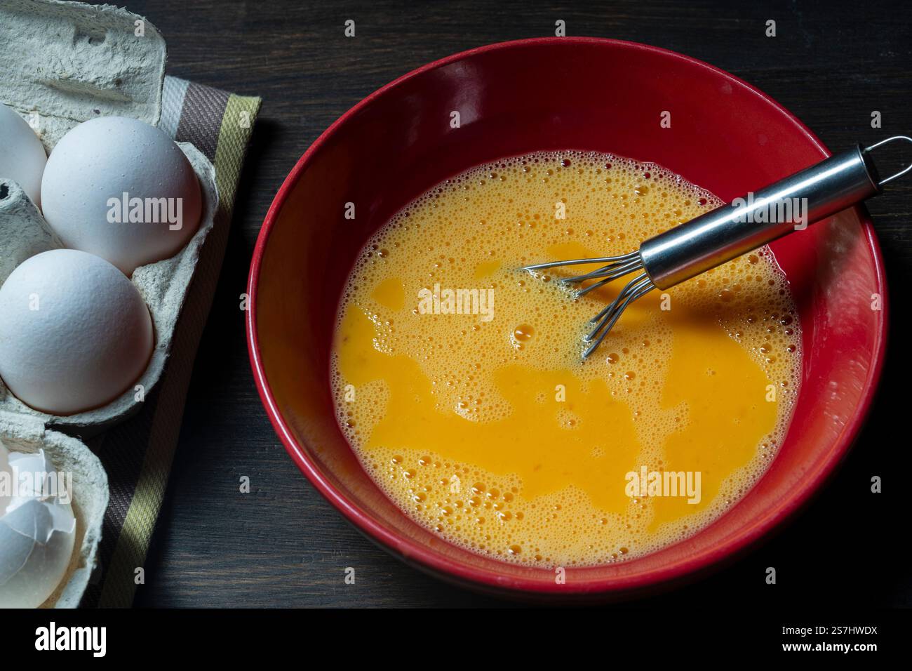 Eier rühren. Rohe Eier in einer roten Schüssel mit einem Schneebesen für Omelette geschlagen, Nahaufnahme. Frühstückszubereitung, rustikaler Stil Stockfoto