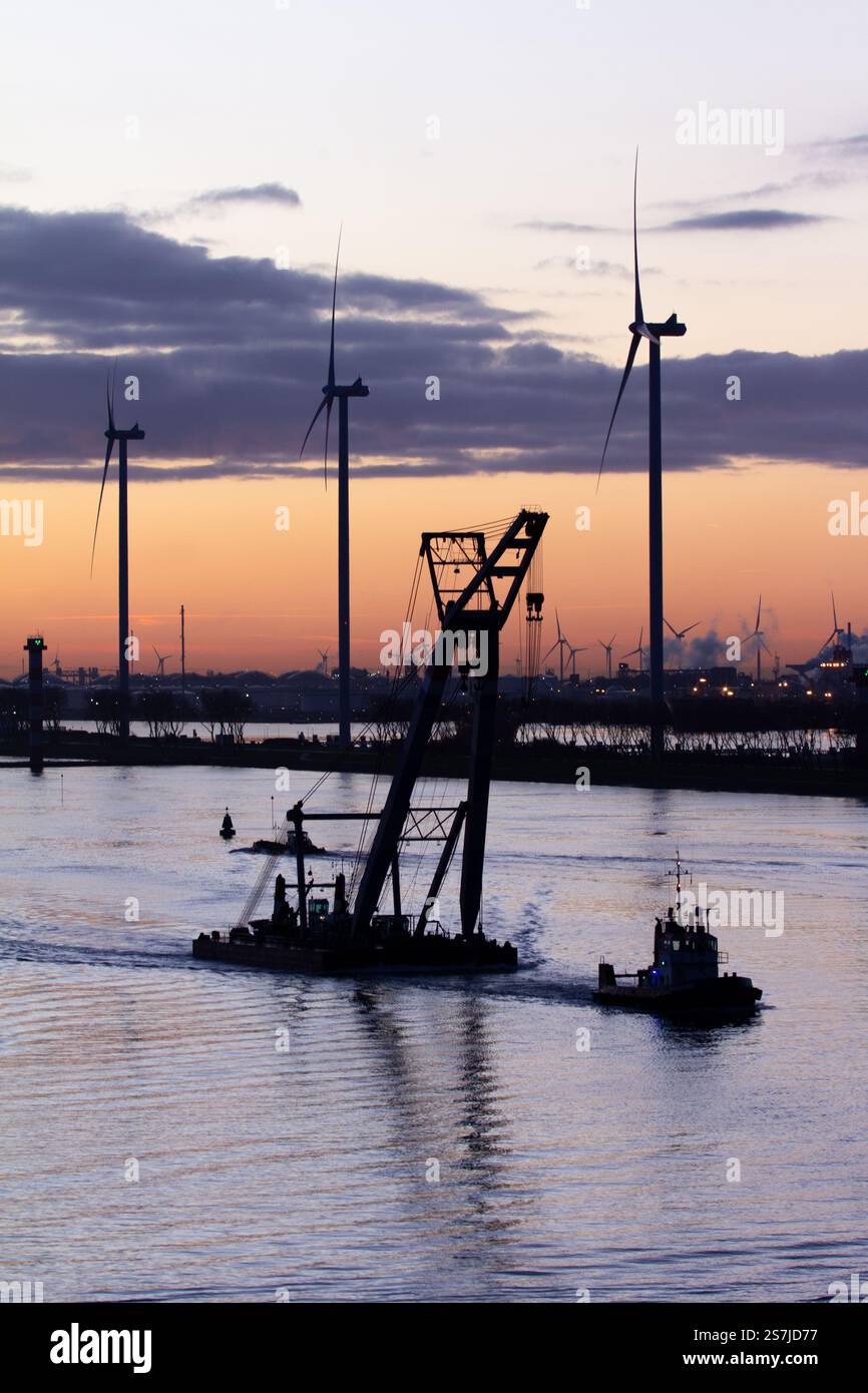 Ein Tug-Boot, das bei Sonnenaufgang in den Niederlanden eine schwimmende Kranplattform schleppt, mit Windturbinen im Hintergrund Stockfoto