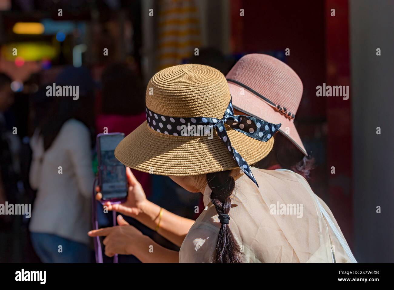 Zwei chinesische (asiatische) Frauen mit breiter Krempe, bedeckten Gesichtern und einem Mobiltelefon bei starkem Sonnenlicht Stockfoto