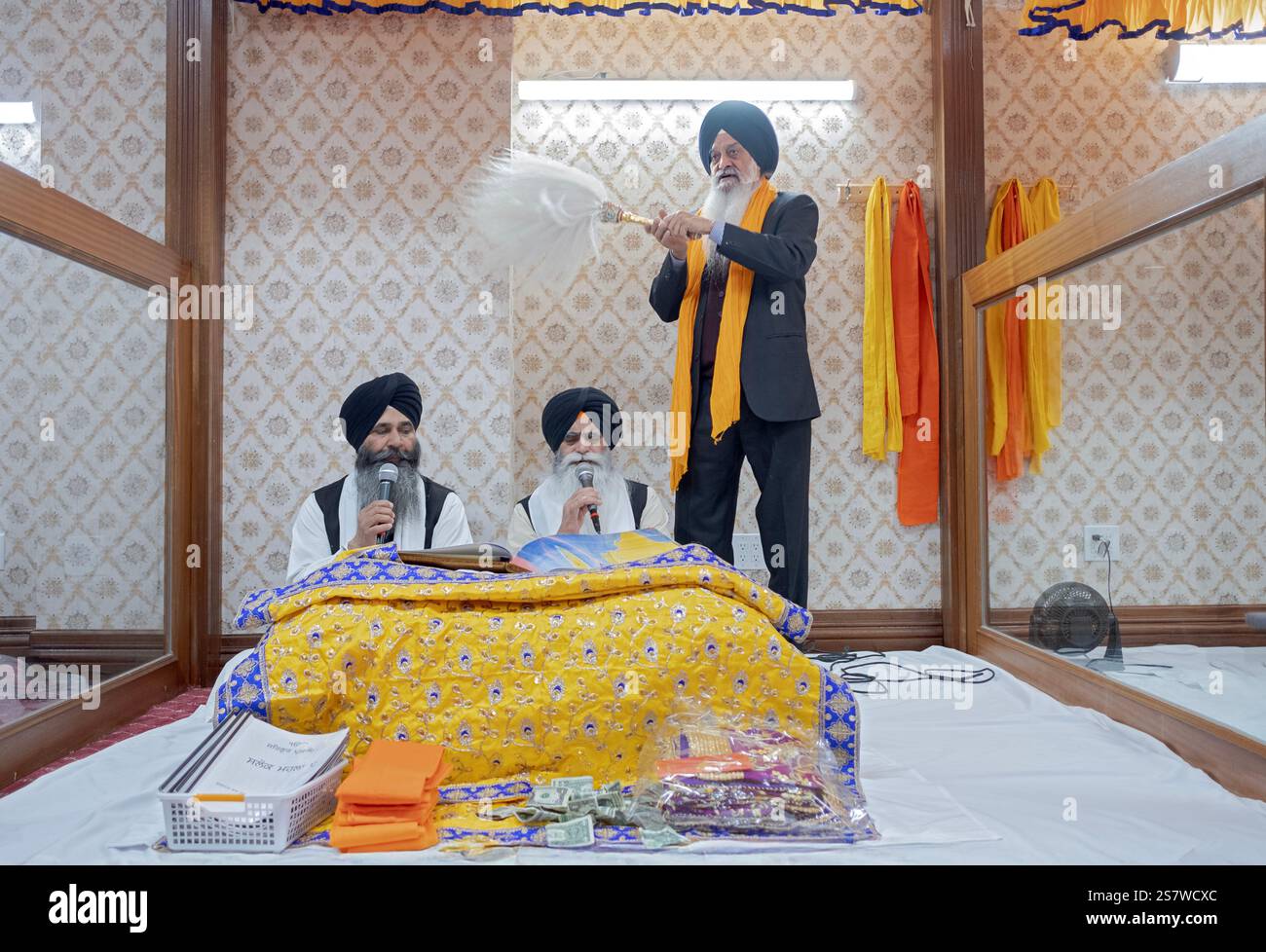 Bei einem Morgengottesdienst in einem Sikh-Tempel lasen Priester ihr heiliges Buch Guru Granth Shaib, während ein dritter Mann respektvoll einen weißen Chaur Sahib winkt. In Queens. Stockfoto
