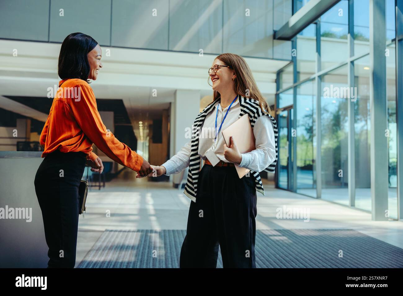 Zwei professionelle Frauen, die einen warmen Handschlag an einem Büroeingang austauschen, repräsentieren ein einladendes Geschäftsumfeld und Karrieremöglichkeiten. Stockfoto