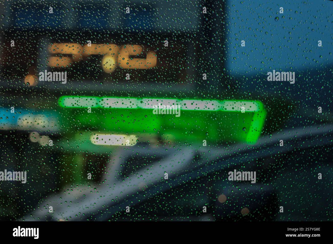 Ein regnerischer Stadtabend durch ein Glasfenster, mit scharfen Regentropfen im Vordergrund und einem verschwommenen Hintergrund mit leuchtenden Neonlichtern in Farbtönen Stockfoto