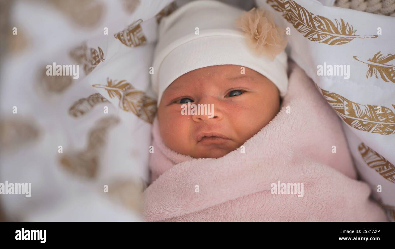Das Neugeborene trägt einen weißen Hut mit einer kleinen Blumendekoration, in eine rosa Decke gewickelt und liegt in einer Krippe mit Federmustern Stockfoto