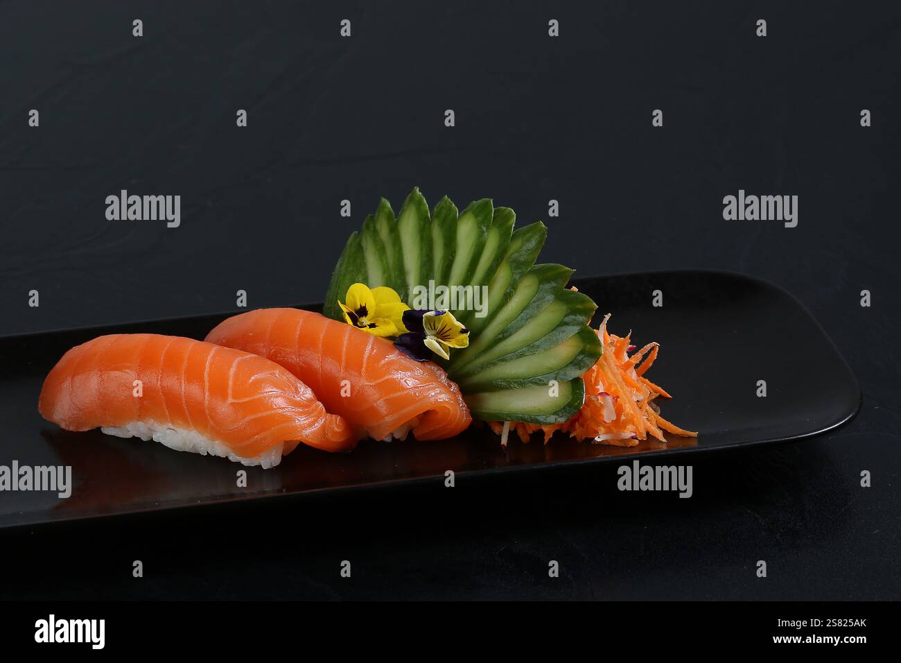 Zwei Stücke Lachs-Nigiri-Sushi, serviert auf einem schwarzen Teller, garniert mit Gurkenscheiben, essbaren Blumen und geriebenem Gemüse. Stockfoto