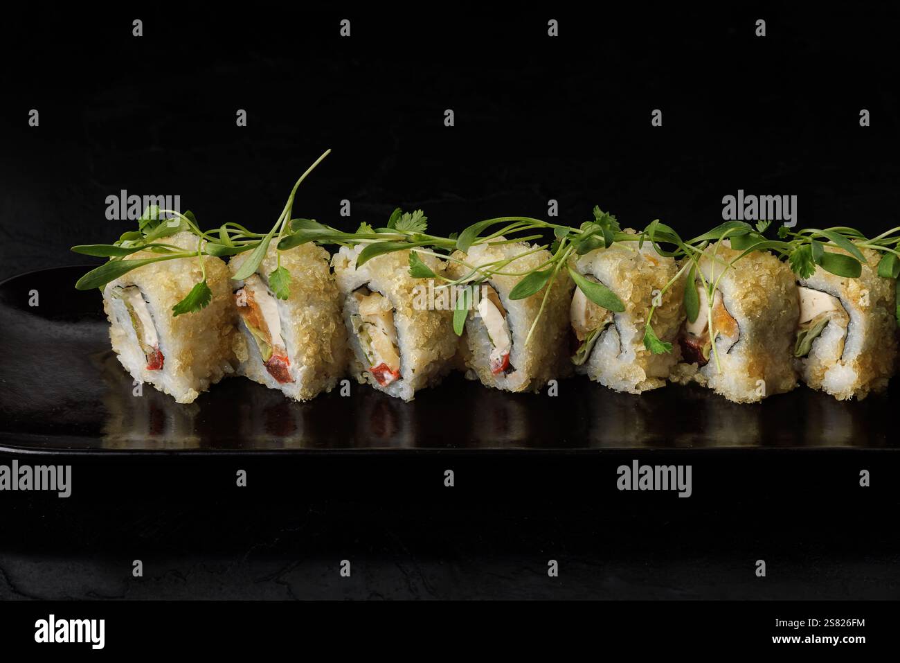 Eine Reihe goldener Tempura-Sushi-Brötchen, garniert mit frischem grünem Mikrogrün, elegant auf einem dunklen rechteckigen Teller angeordnet. Die Rollen sind knusprig Stockfoto