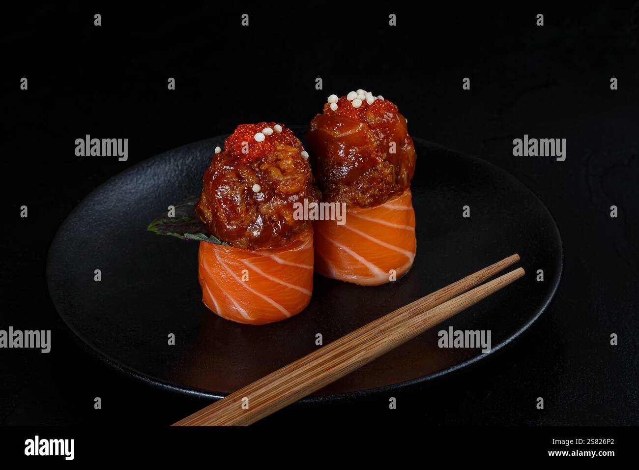 Zwei Stücke Lachs-Sushi umwickelt um scharfe Toppings, garniert mit Fischrogen und knusprigen Perlen, elegant präsentiert auf einem schwarzen Teller mit Chopstik Stockfoto