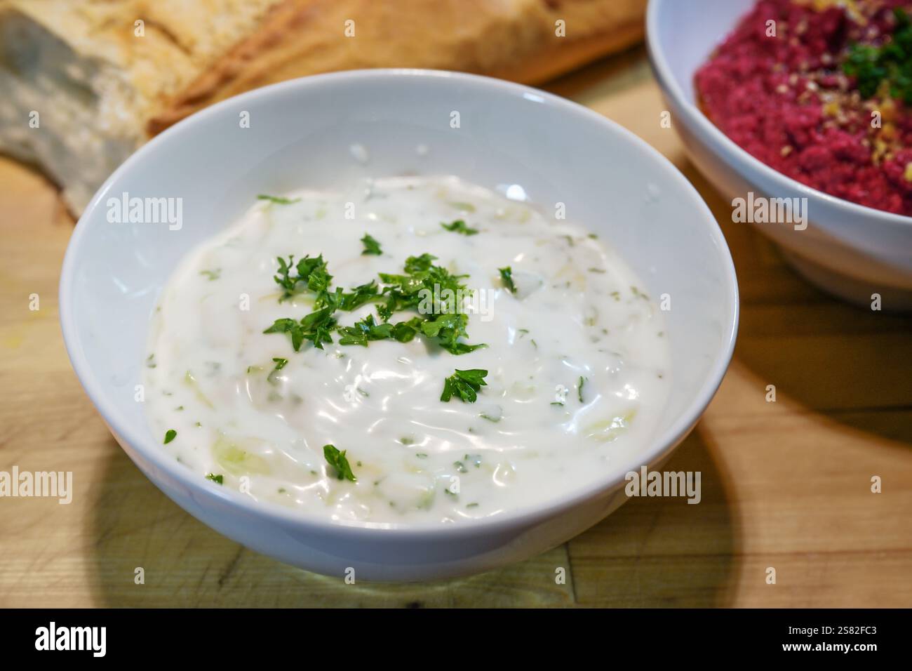 Gesunder Dip aus veganem Joghurt mit Gurke, Knoblauch und Kräutern mit Petersilie garniert in einer weißen Schüssel mit Brot auf rustikalem Holzbrett, hausgemacht Stockfoto