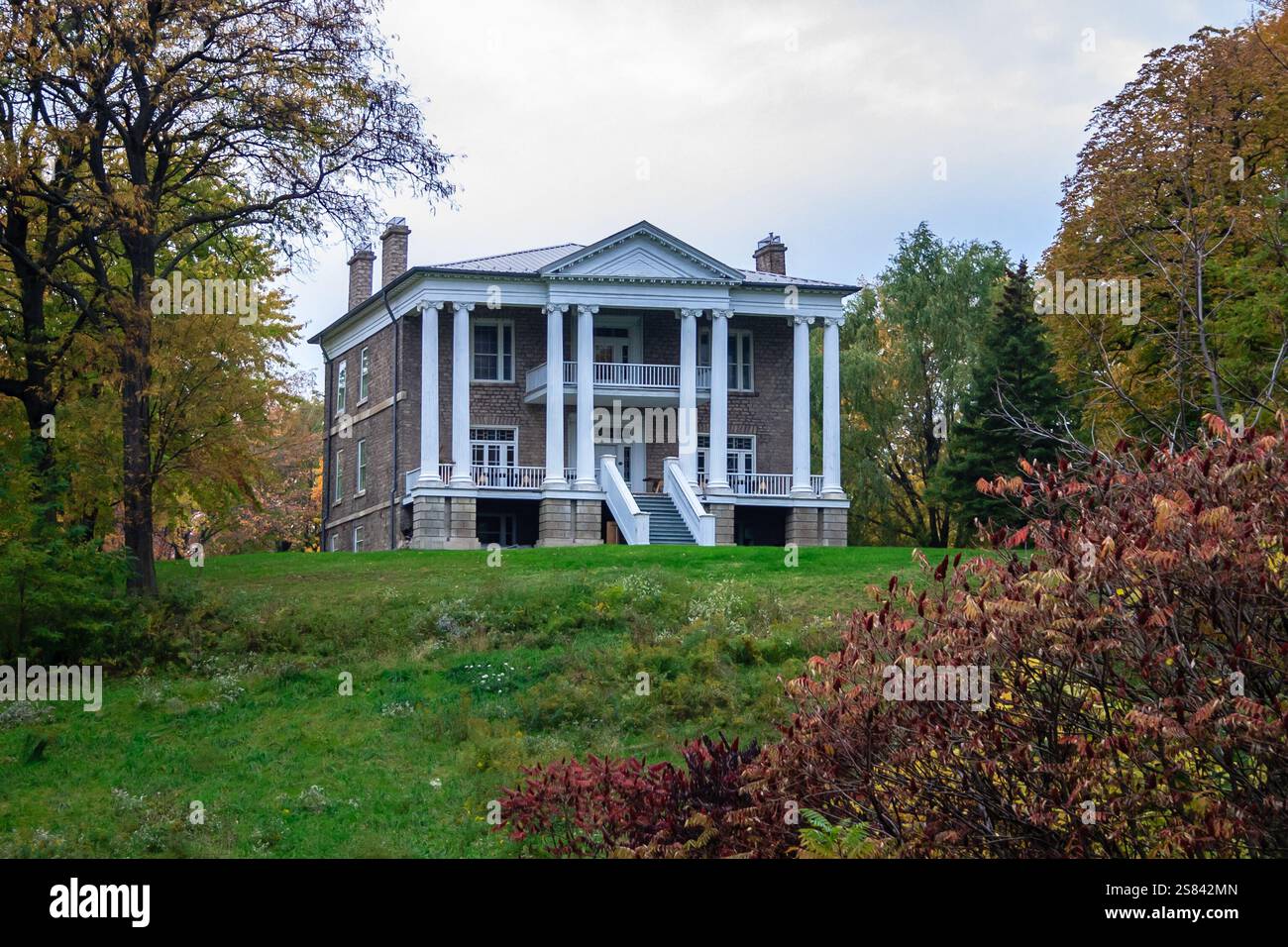 Ein großes Haus mit weißer Fassade liegt auf einem Hügel. Das Haus ist von Bäumen und Büschen umgeben und das Gras ist üppig und grün. Das Haus hat einen Riesen Stockfoto