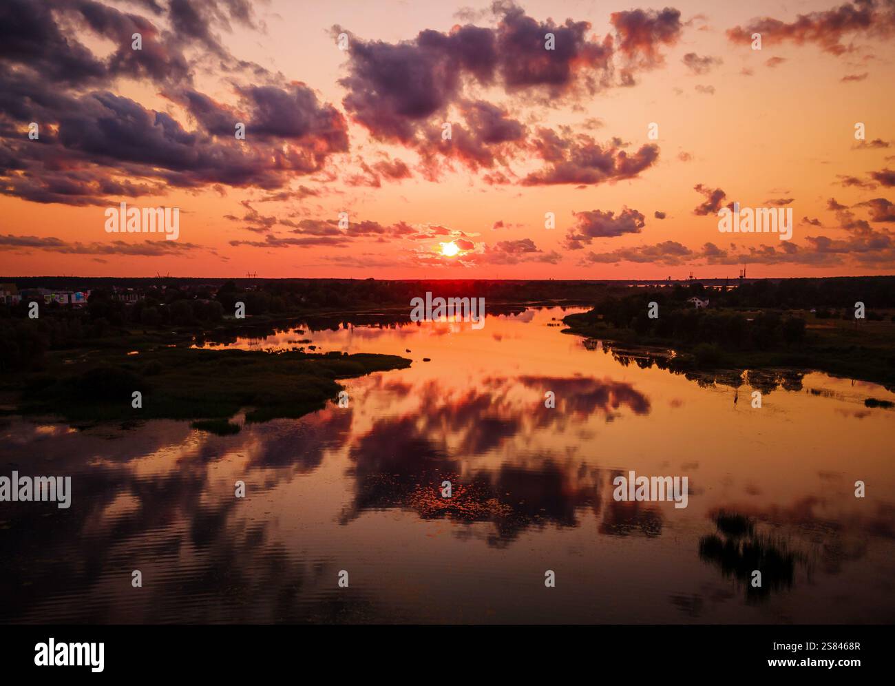 Eine niedrige Sonne wirft Orange- und Rottöne über den Himmel, die sich in einem ruhigen Fluss spiegeln. Vegetation und entfernte Baumsilhouetten verleihen der Szene Tiefe. Stockfoto