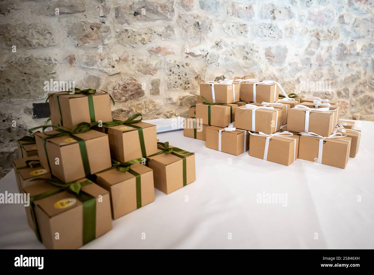Zwei Stapel kleiner brauner Geschenkboxen, die mit grünen und weißen Bändern gebunden sind, sitzen auf einer weißen Tischdecke vor einer strukturierten Steinmauer. Stockfoto