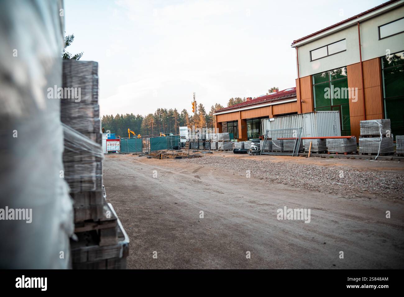 Teilweise fertiggestelltes Gebäude mit grün getönten Fenstern und rotem Dach, umgeben von Baumaterialien, Ausrüstung und einer bewaldeten Kulisse. Stockfoto