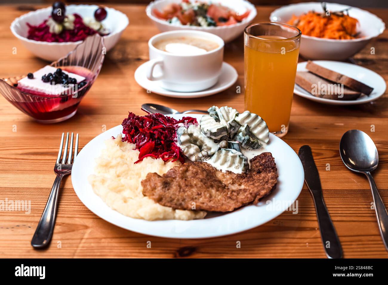 Ein Holztisch zeigt eine Mahlzeit mit Kartoffelpüree, paniertem Fleisch, Rotkohl, Gurkensalat, Beilagen, Dessert, Orangensaft und Kaffee. Stockfoto