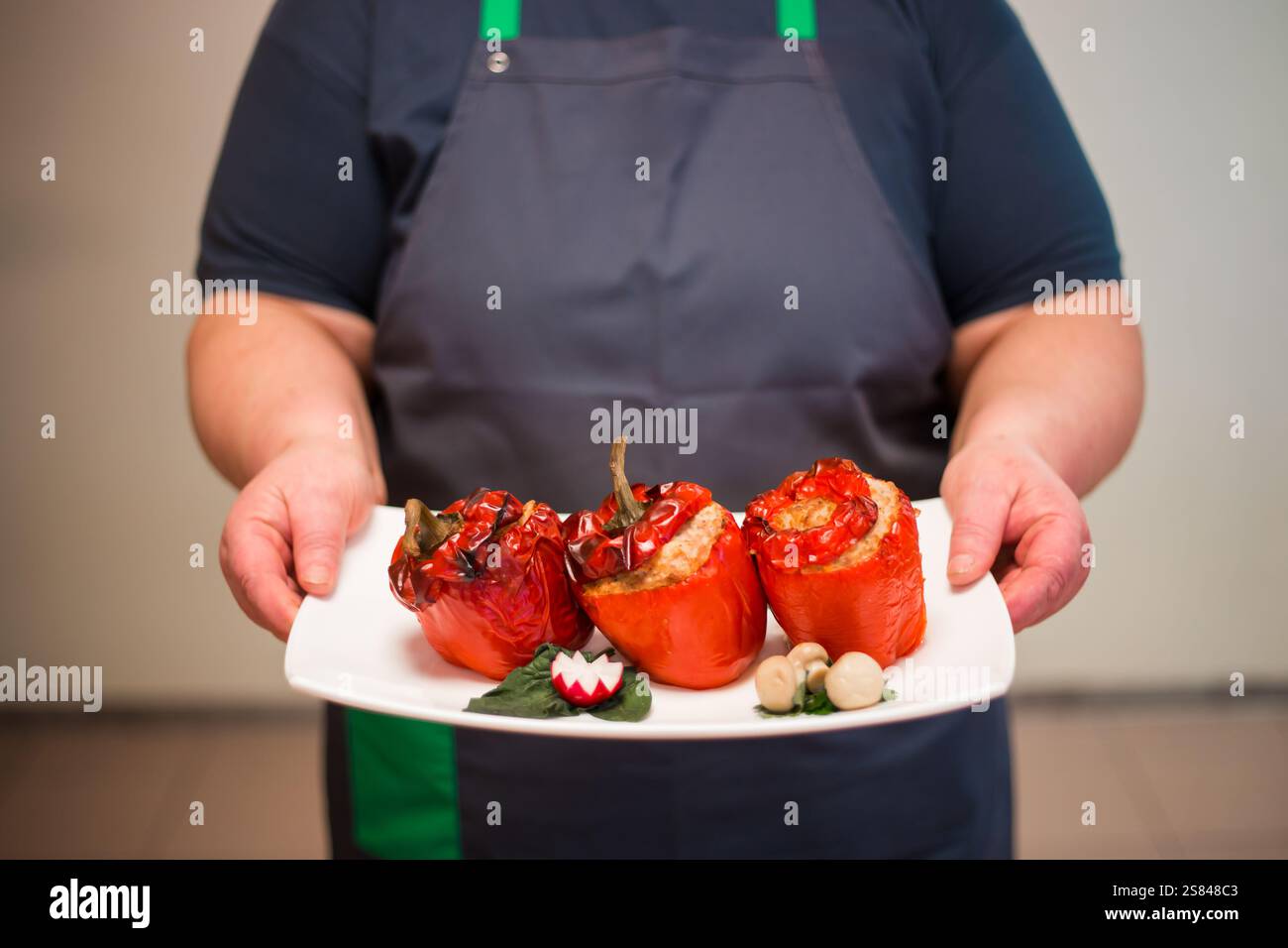 Eine Person in einer dunklen Schürze hält einen weißen Teller mit gerösteten roten Paprika, gefüllt mit Körnern, garniert mit einem geschnitzten Rettich und eingelegten Zwiebeln. Stockfoto