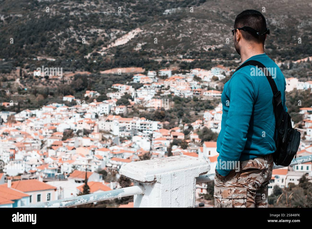 Ein Mann in blauem Hemd und Tarnhosen steht auf einem weißen Balkon mit schwarzem Rucksack und beobachtet eine Stadt mit weißen Gebäuden und zerklüfteten Hügeln dahinter. Stockfoto
