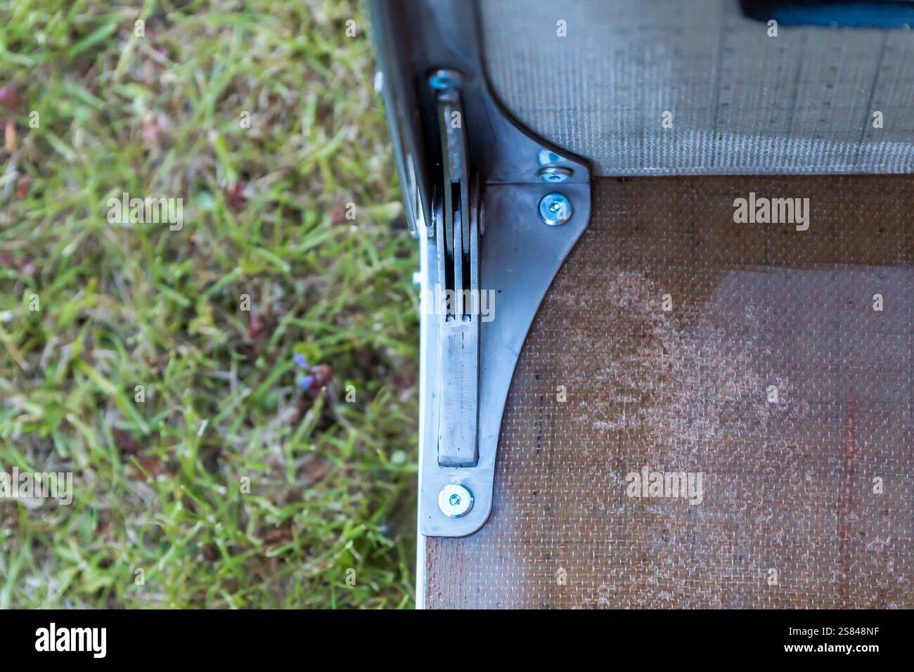 Detaillierte Ansicht eines Metallscharniers, das mit Schrauben auf einer Holzoberfläche befestigt ist und mit kleinen roten Elementen im Freien gegen Gras gestellt wird. Stockfoto