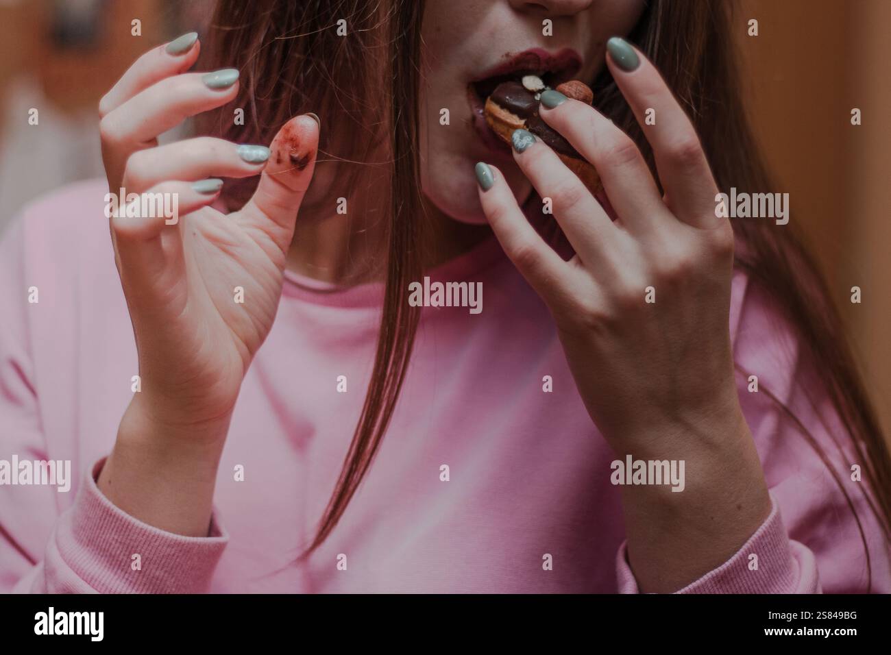 Eine Person in einem rosafarbenen Sweatshirt isst einen Schokoladen-Macaron, mit Schokoladenflecken auf Lippen und Fingern. Grünlicher nagellack und verschwommener warmer Hintergrund V Stockfoto
