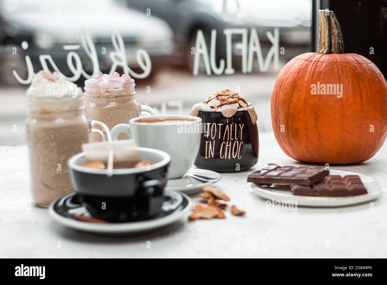 Ein gemütlicher Kaffeetisch bietet Tassen mit heißen Getränken, Kürbis, Schokoladentafeln und Keksen. Ein Fenster mit handgeschriebenem Text und unscharfen Autos sind sichtbar Stockfoto
