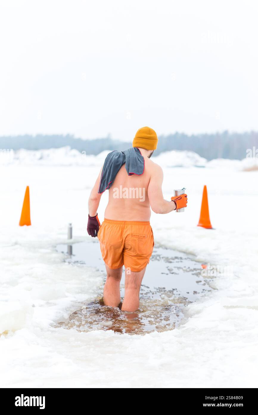 Ein Individuum in orangefarbener Kleidung waten durch ein eingeschnittenes Loch in ein gefrorenes Gewässer, umgeben von Schnee, Verkehrskegeln und einer bewaldeten Winterkulisse. Stockfoto
