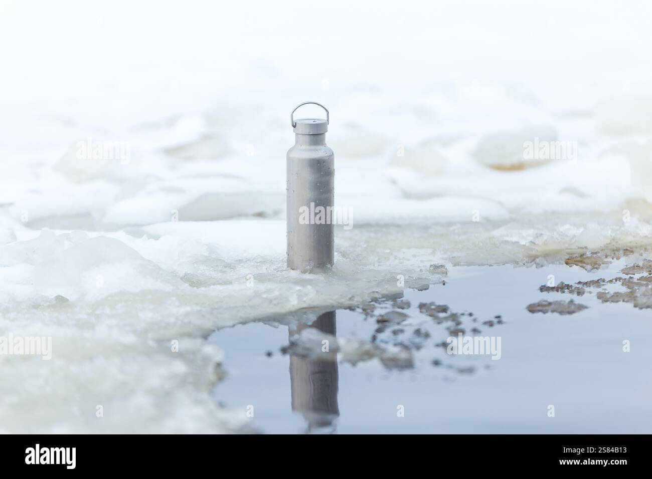 Eine metallische Wasserflasche steht auf Eis neben einem teilweise gefrorenen Wasserkörper. Schnee und Eis bedecken den Bereich mit sanfter Beleuchtung und einem ruhigen Reflektionsvisier Stockfoto