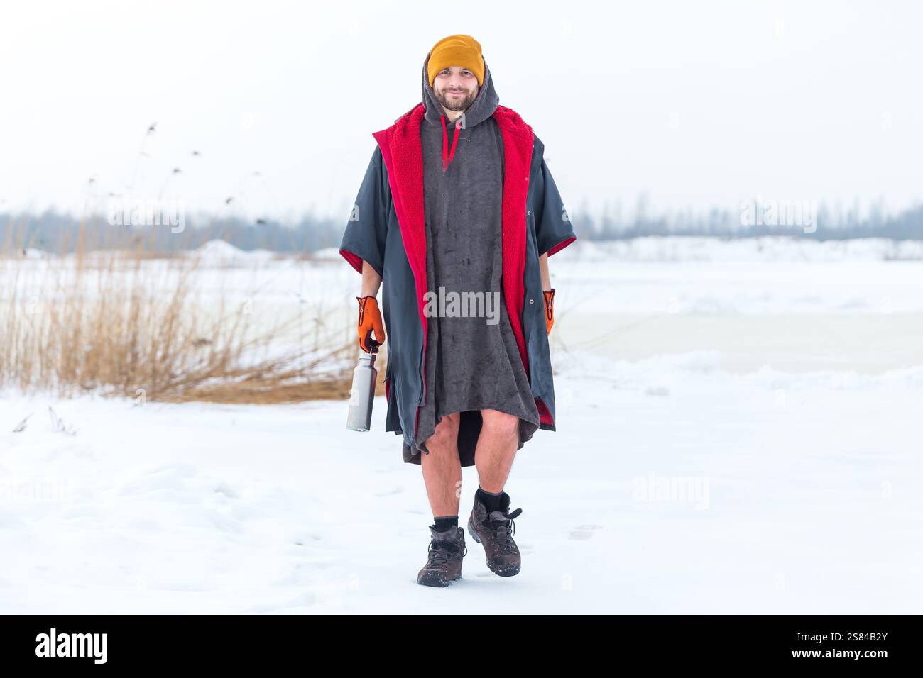 Ein Mann in einem Kapuzenmantel mit rotem Innenraum steht auf einem verschneiten Gelände und hält eine Wasserflasche aus Metall. Spärliches Schilf, gefrorenes Wasser und Bäume im Ba Stockfoto