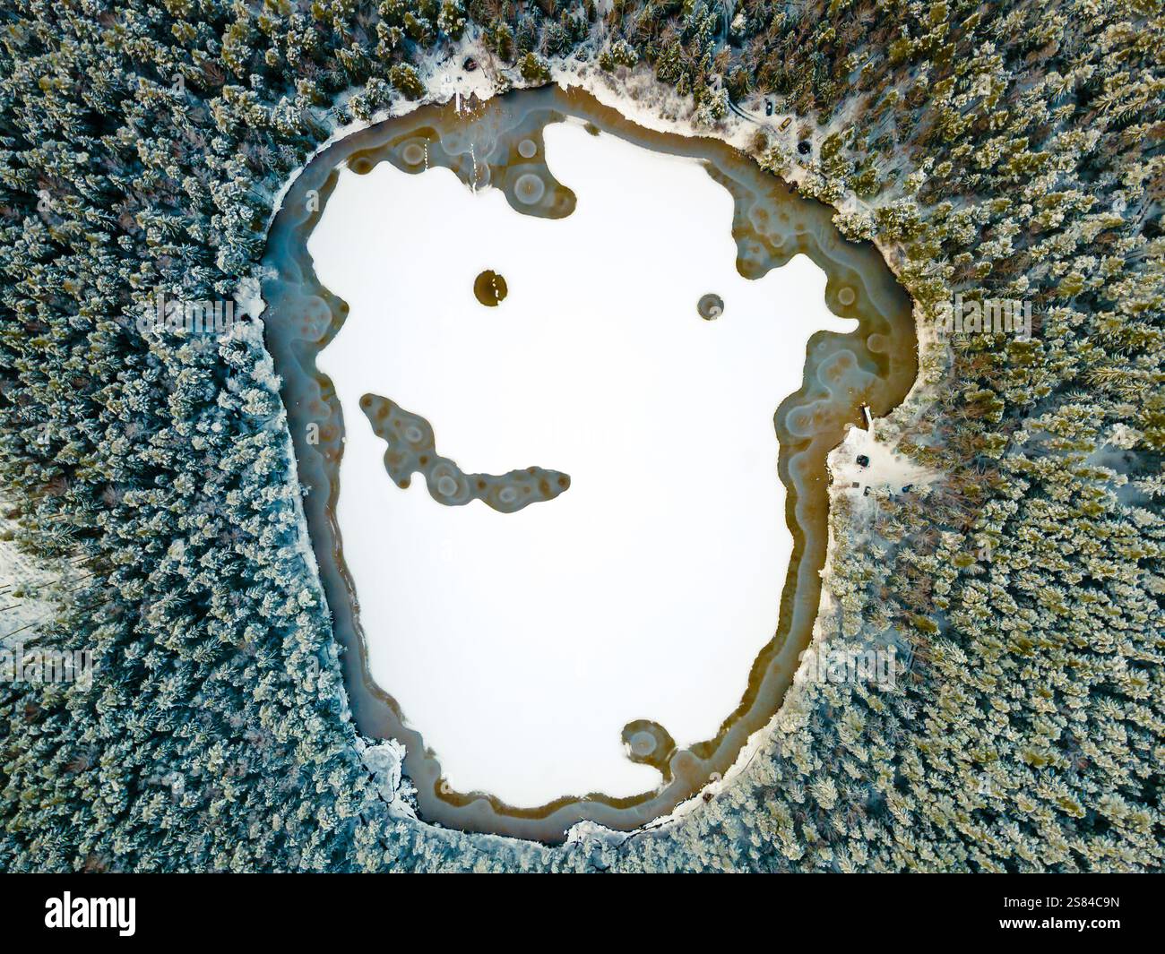 Gefrorener See mit einzigartigen Eismustern, die einem lächelnden Gesicht ähneln, umgeben von einem dichten Wald aus schneebedeckten Bäumen, die sich symmetrisch nach außen erstrecken. Stockfoto