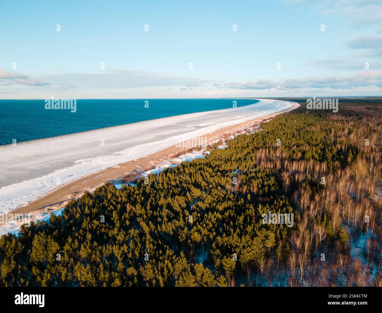 Eine gefrorene Küste trifft auf ein riesiges blaues Meer, umgeben von einem dichten immergrünen Wald. Schnee bedeckt Teile des Bodens unter klarem Himmel mit verstreuten Wolken Stockfoto
