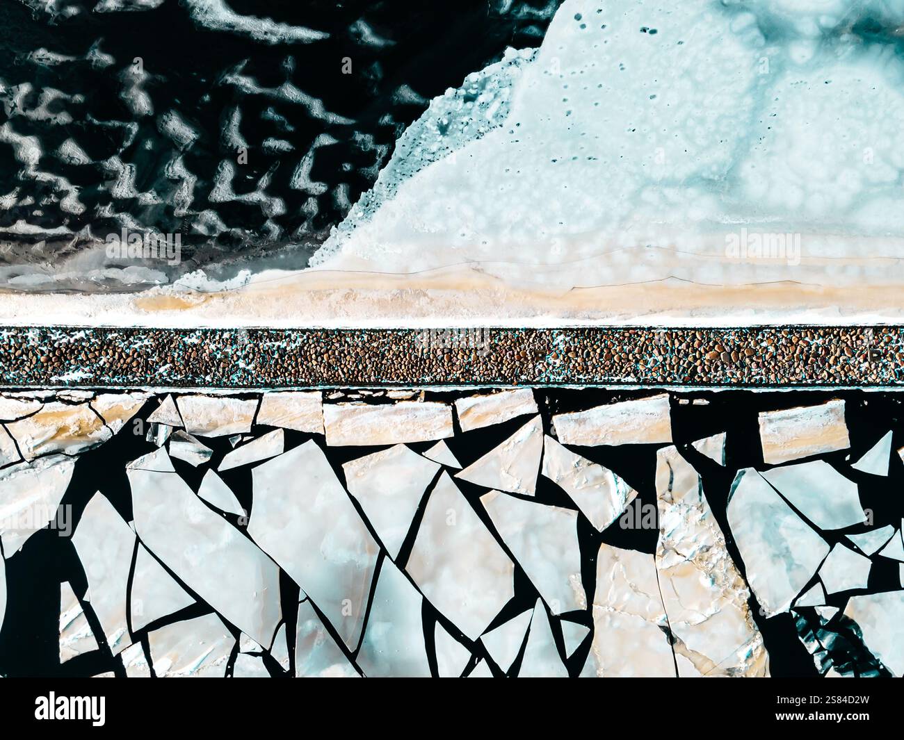 Eckige Eisschilde treffen auf welliges dunkles Wasser, getrennt durch einen schmalen, mit Kies bedeckten Landstreifen, möglicherweise eine Straße oder ein Damm, in dieser Luftaufnahme. Stockfoto