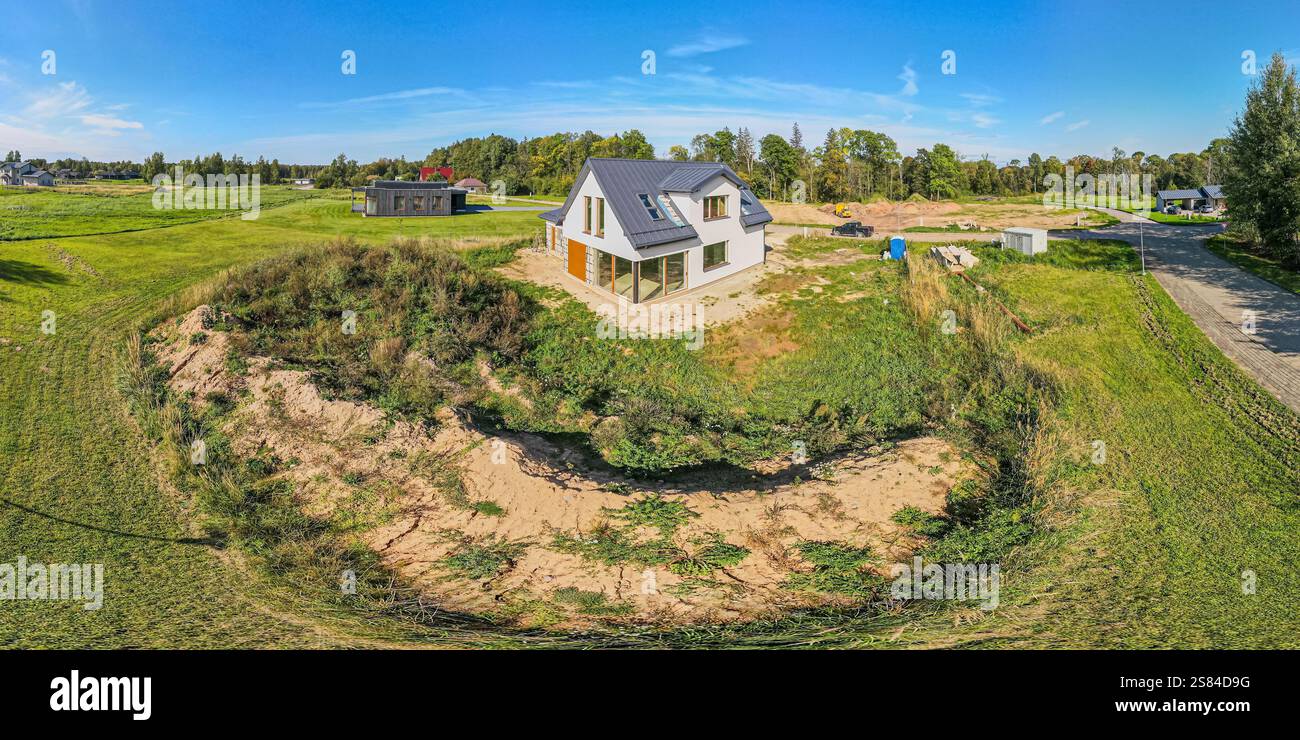 Ein modernes zweistöckiges Haus mit großen Fenstern und einem dunklen Dach, umgeben von Gras und karger Vegetation, mit einer sandigen Vertiefung im Vordergrund. Stockfoto