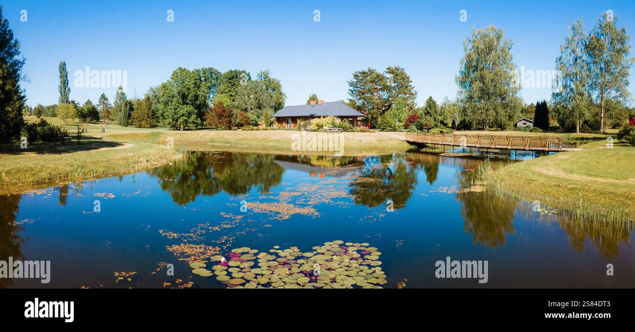 Ein Teich mit Seerosenpolstern spiegelt den blauen Himmel und die Bäume wider, mit einer hölzernen Fußbrücke, die zu einem Haus mit dunklem Dach und Kamin führt, umgeben von La Stockfoto