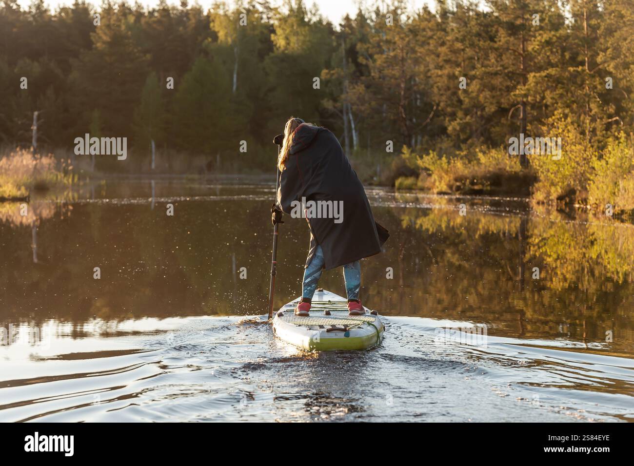 Eine Person in einem dunklen Mantel und Jeans-Paddleboards auf ruhigem Wasser, umgeben von immergrünen und Laubbäumen, die sich im warmen Sonnenlicht spiegeln. Stockfoto