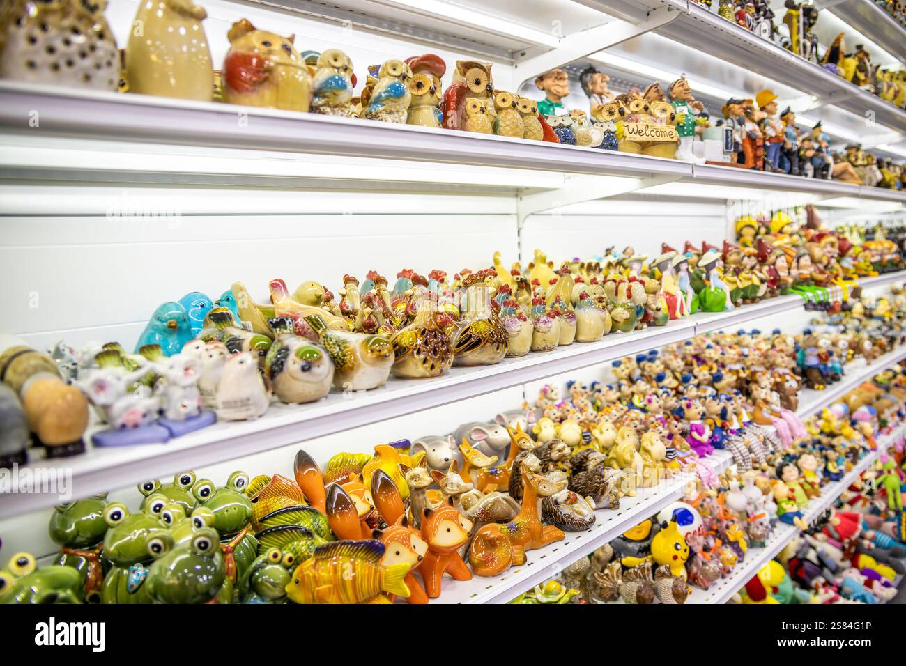 Ordentlich angeordnete Keramik- und Glasfiguren, darunter Frösche, Eulen und Vögel, auf weißen Regalen unter heller Beleuchtung mit einem hölzernen Willkommensschild. Stockfoto