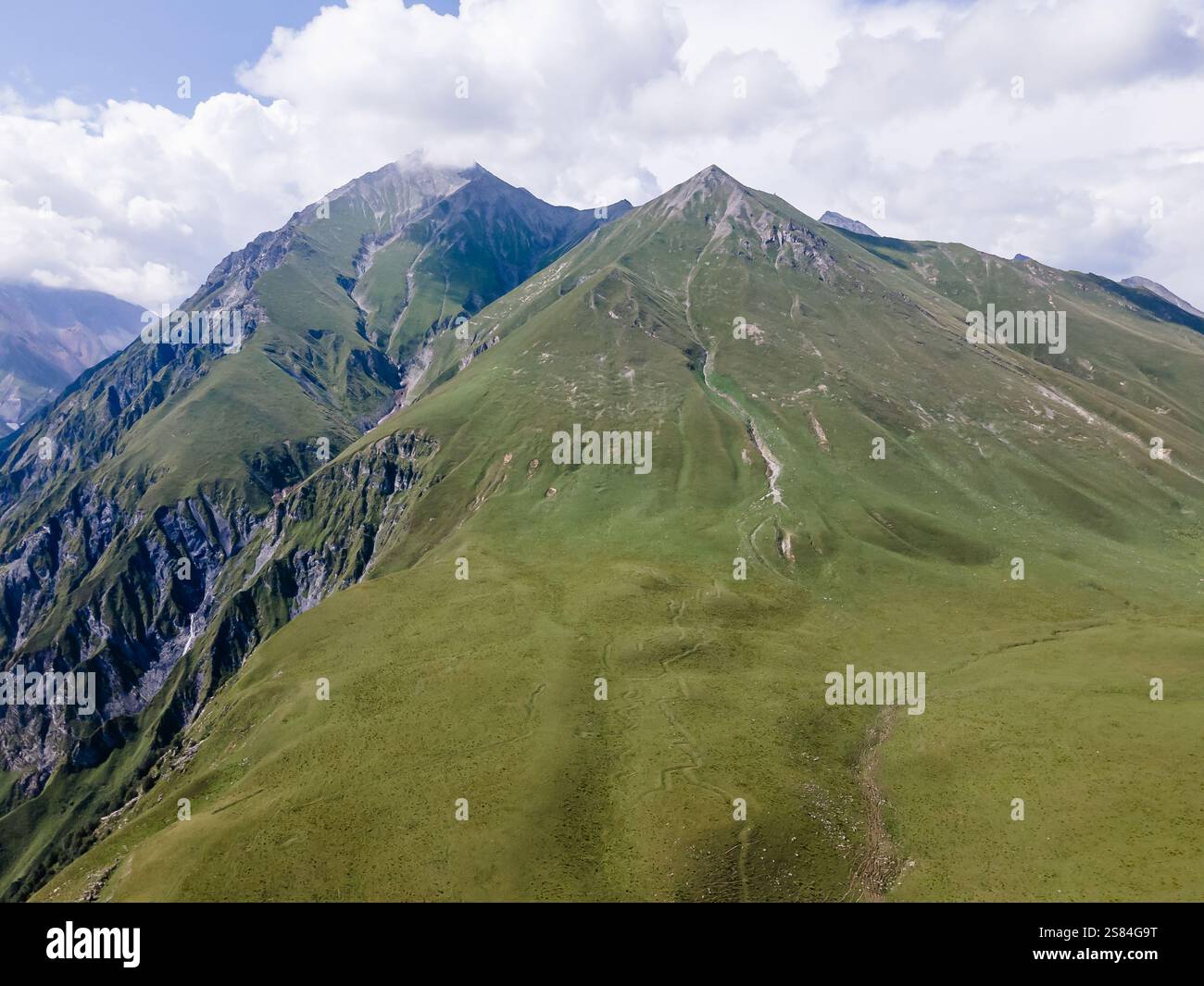 Steile Berghänge mit grasbewachsenen Gipfeln und freiliegendem Fels, durchzogen von sichtbaren Pfaden unter teilweise bewölktem Himmel, wahrscheinlich in Georgia. Stockfoto
