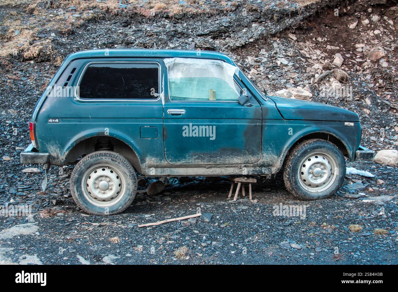 Ein verwitterter blauer Lada Niva 4x4 ruht auf unebenem, felsigem Boden, der für Reparaturen abgestellt wird. Die Kulisse umfasst einen kargen Hügel mit karger Vegetation. Stockfoto
