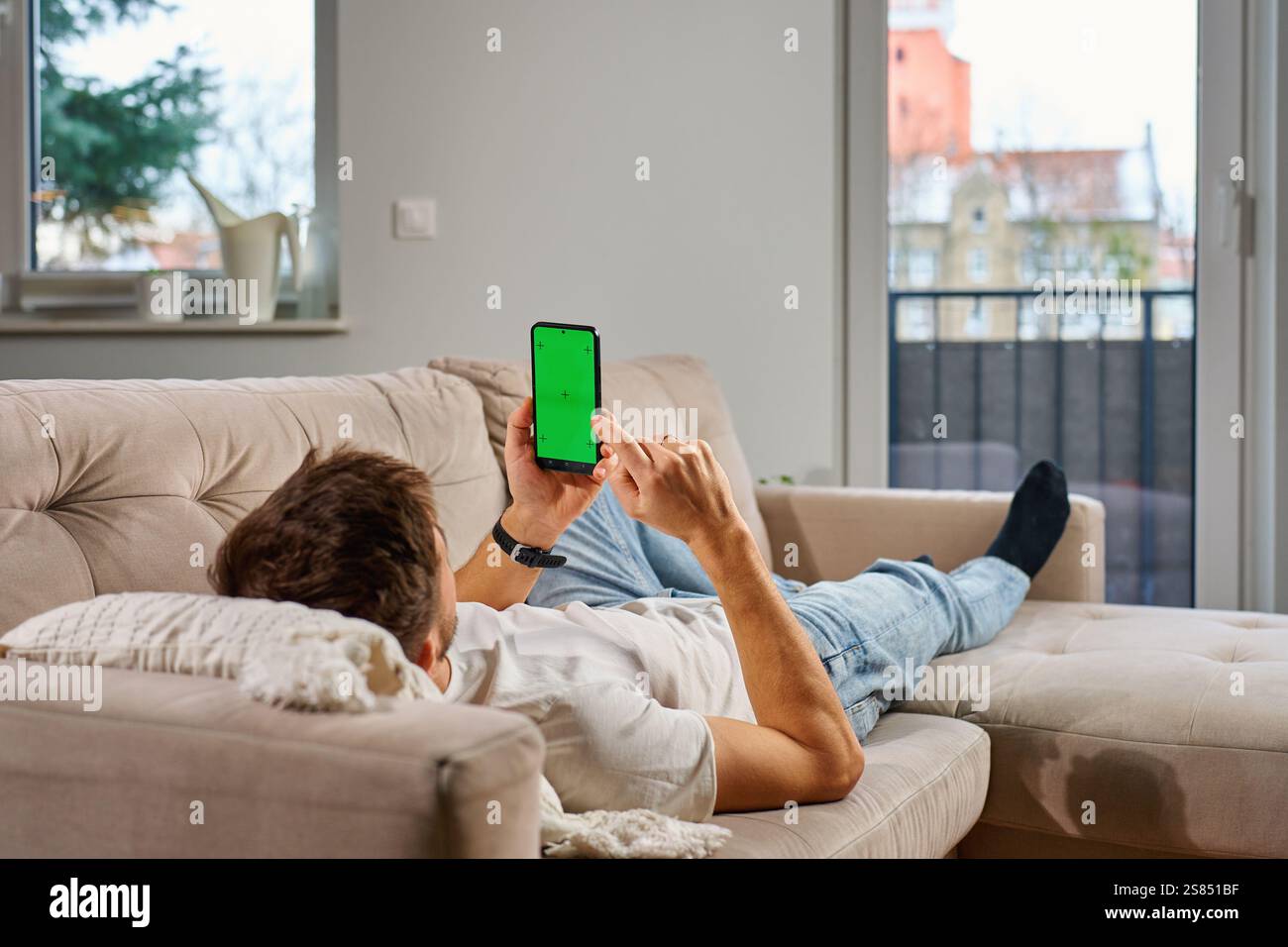 Ein Mann in lässiger Kleidung liegt auf der beigen Couch, hält ein Smartphone mit grünem Bildschirm und entspannt sich im modernen Wohnzimmer. Mann, der sich auf dem Sofa ausruht und sich anschaut Stockfoto
