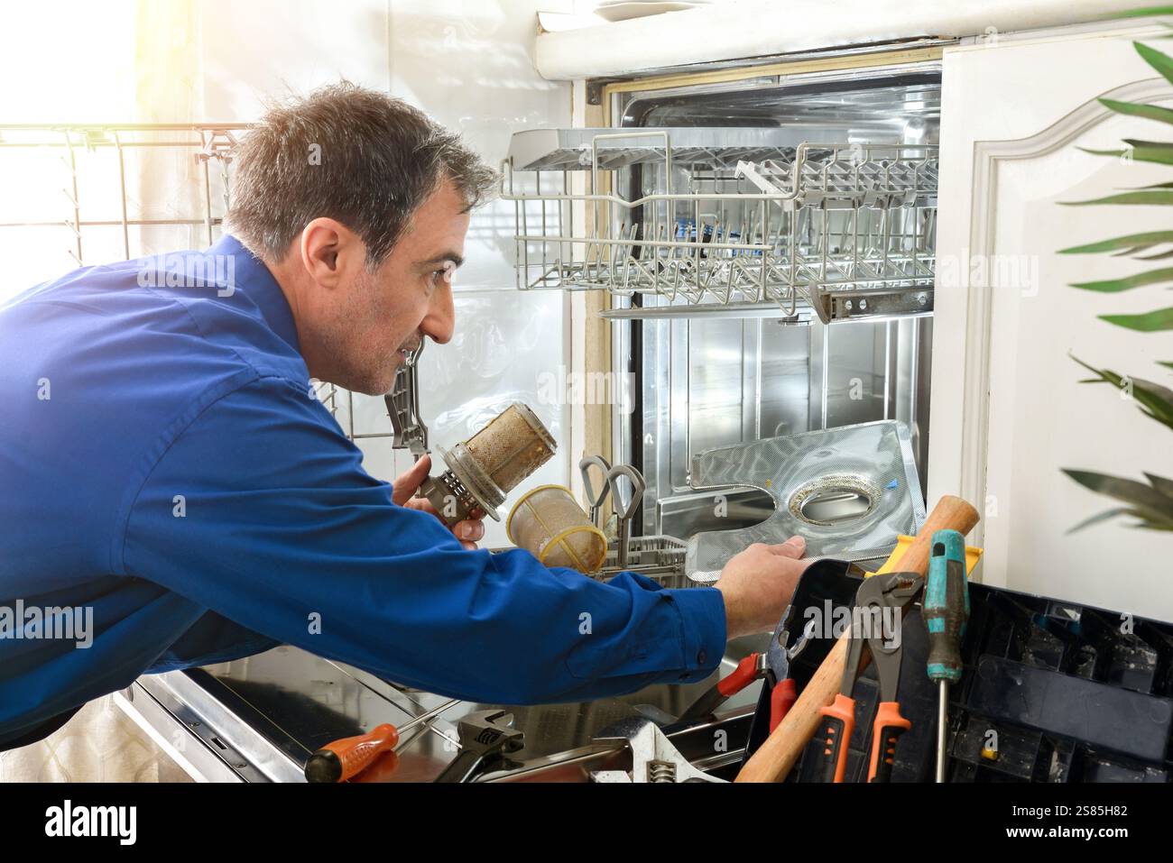 Detail eines männlichen Technikers, der als blauer Arbeiter gekleidet ist und einen Geschirrspüler mit Werkzeugen in einer weißen Küche vorbereitet Stockfoto