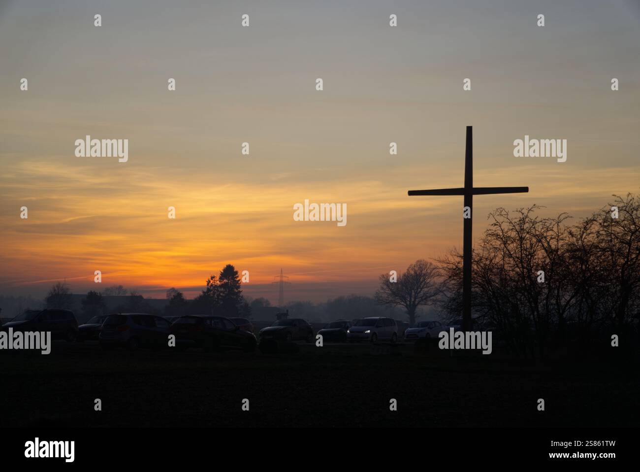 Geparktes Auto mit eingeschalteten Scheinwerfern zwischen unbeleuchteten geparkten Autos unter einem großen Feldkreuz und vor dem gelb-orangen Abendhimmel Stockfoto
