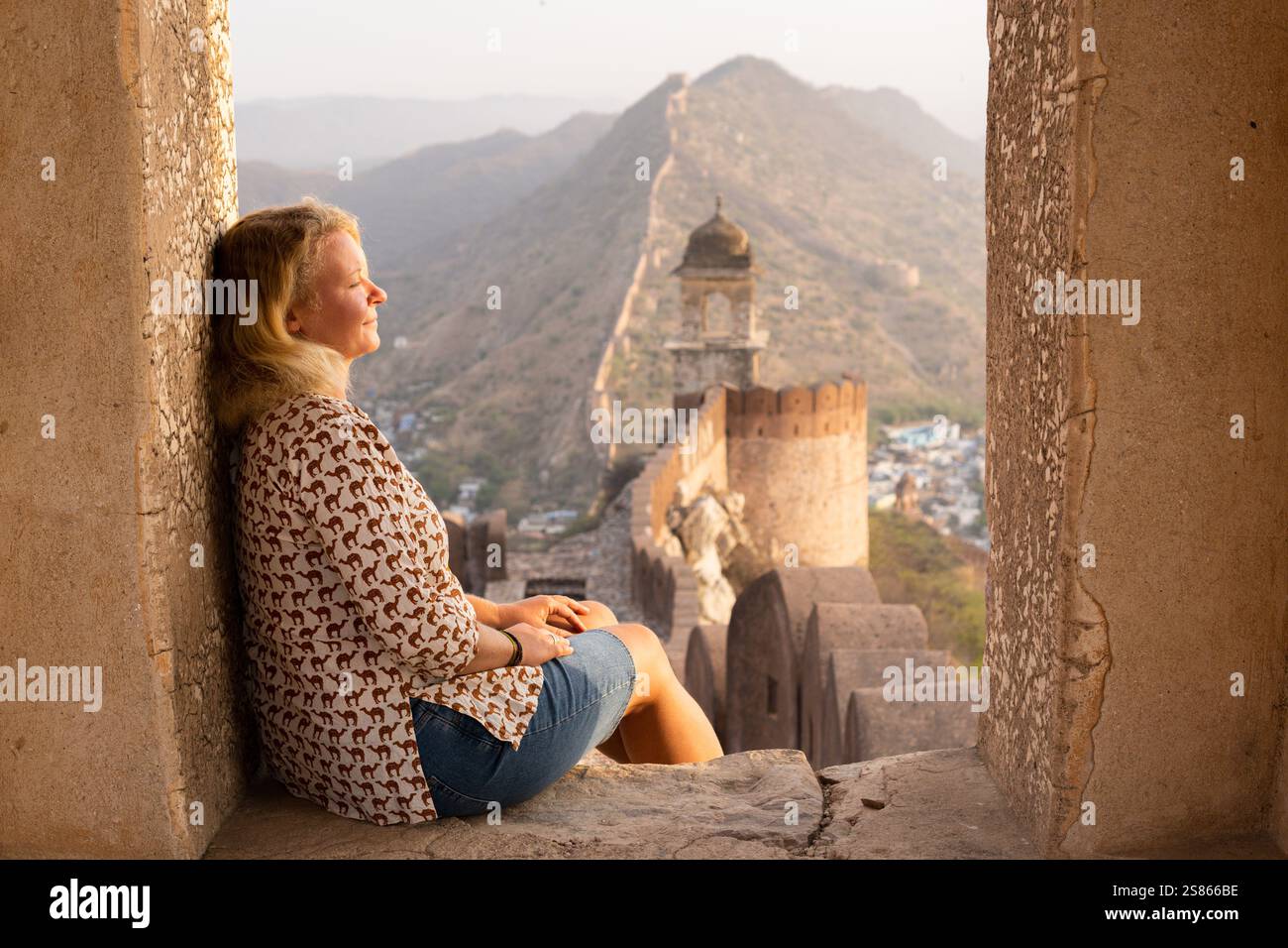 Junge blonde Frau genießt Sonnenaufgang an der Chinesischen Mauer von Jaipur in Rajasthan, Indien. Frühmorgendliches Licht auf den trüben Bergen und Festungsmauern Stockfoto