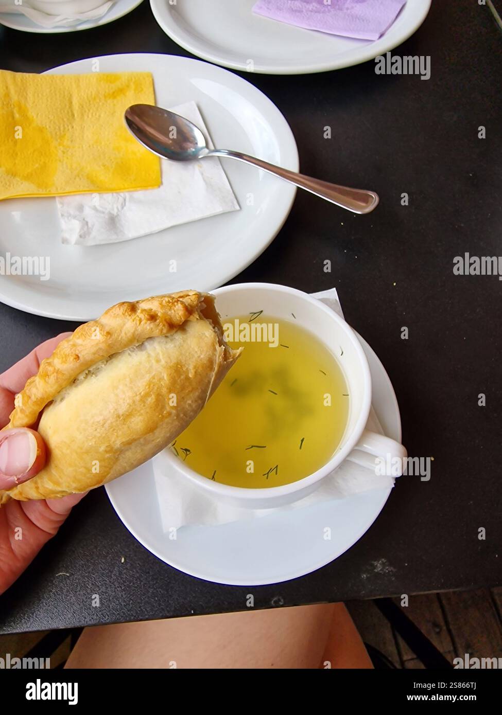 Eine Hand mit einem traditionellen Gebäck (Kibinas) gefüllt mit Hammelfleisch, mit warmer Hühnerbrühe, serviert auf einem dunklen Holztisch, Litauen Stockfoto