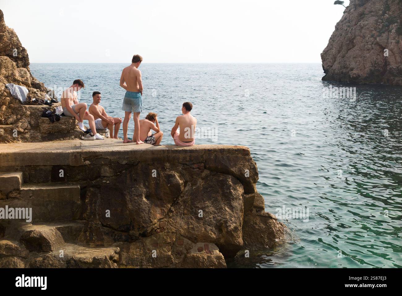 Junge Männer / Reisende im Urlaub / entspannen Sie sich in der Sonne auf Felsen und Betonplattform mit Blick auf das Adriatische Meer am steinigen Strand Sulic, Dubrovnik / Šulić (Kolorina) Strand, Dubrovnik. Kroatien. (138) Stockfoto