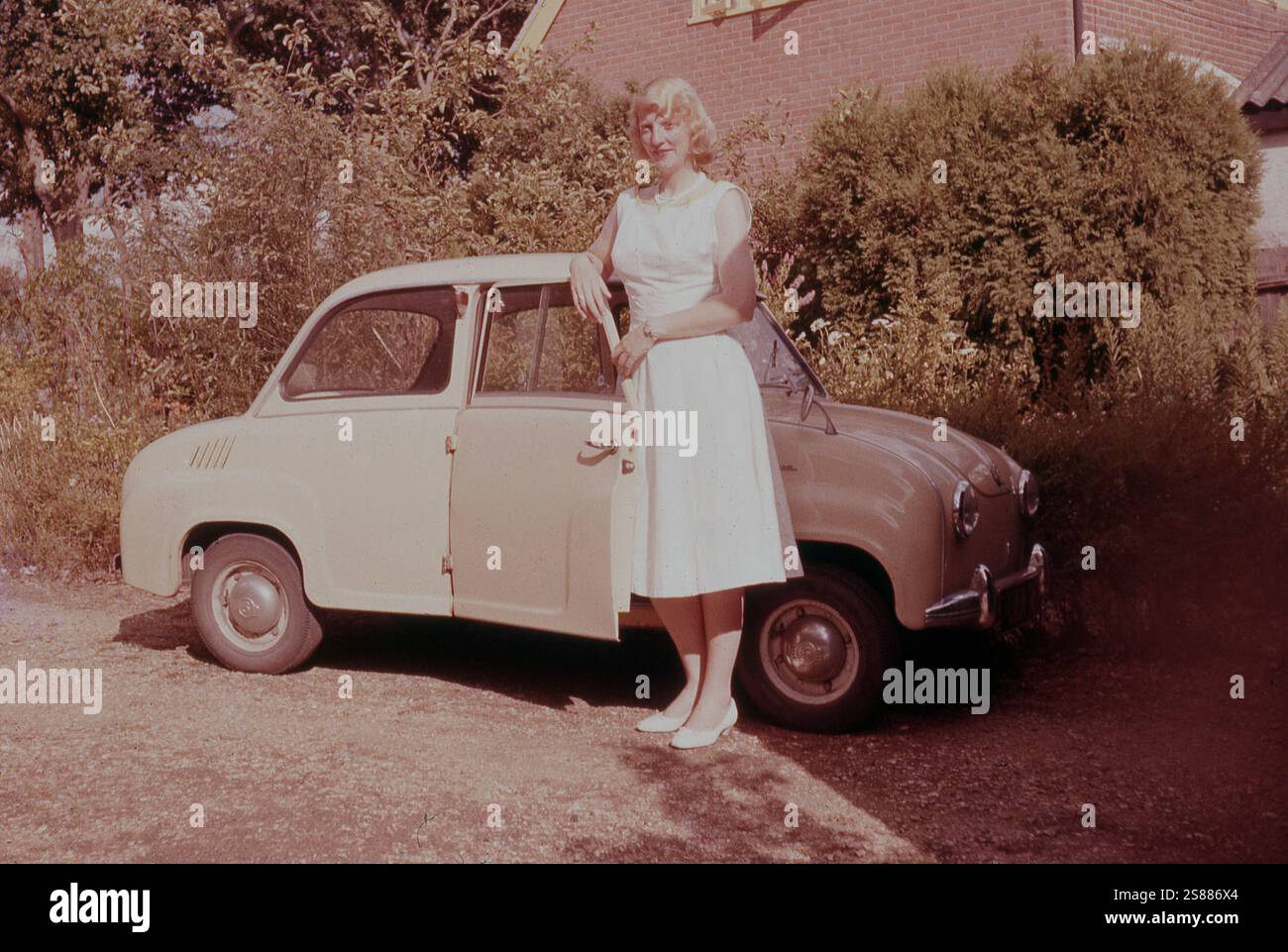1960er Jahre, Sommer und eine junge Frau neben ihrem Goggomobil, einem zweitürigen Kleinwagen mit Heckmotor, hergestellt von Hans Glas in Bayern. Die hier vorgestellte Version, der Glas Goggomobile 250 (T250), hatte ursprünglich nach hinten aufklappbare Türen, ein Merkmal im Fahrzeugdesign, das früher häufig verwendet wurde, aber aus Sicherheitsgründen geändert wurde. Der Motor des Goggomobil wurde 1954 erstmals auf der IFAM Fahrrad- und Motorradmesse gesehen und war ein luftgekühlter Zweitaktmotor mit ursprünglich 250 ccm Hubraum. Diese Kleinwagen, auch bekannt als „Minicars“, wurden in den späten 1940er und 1950er Jahren erstmals als Dreiräder hergestellt. Stockfoto
